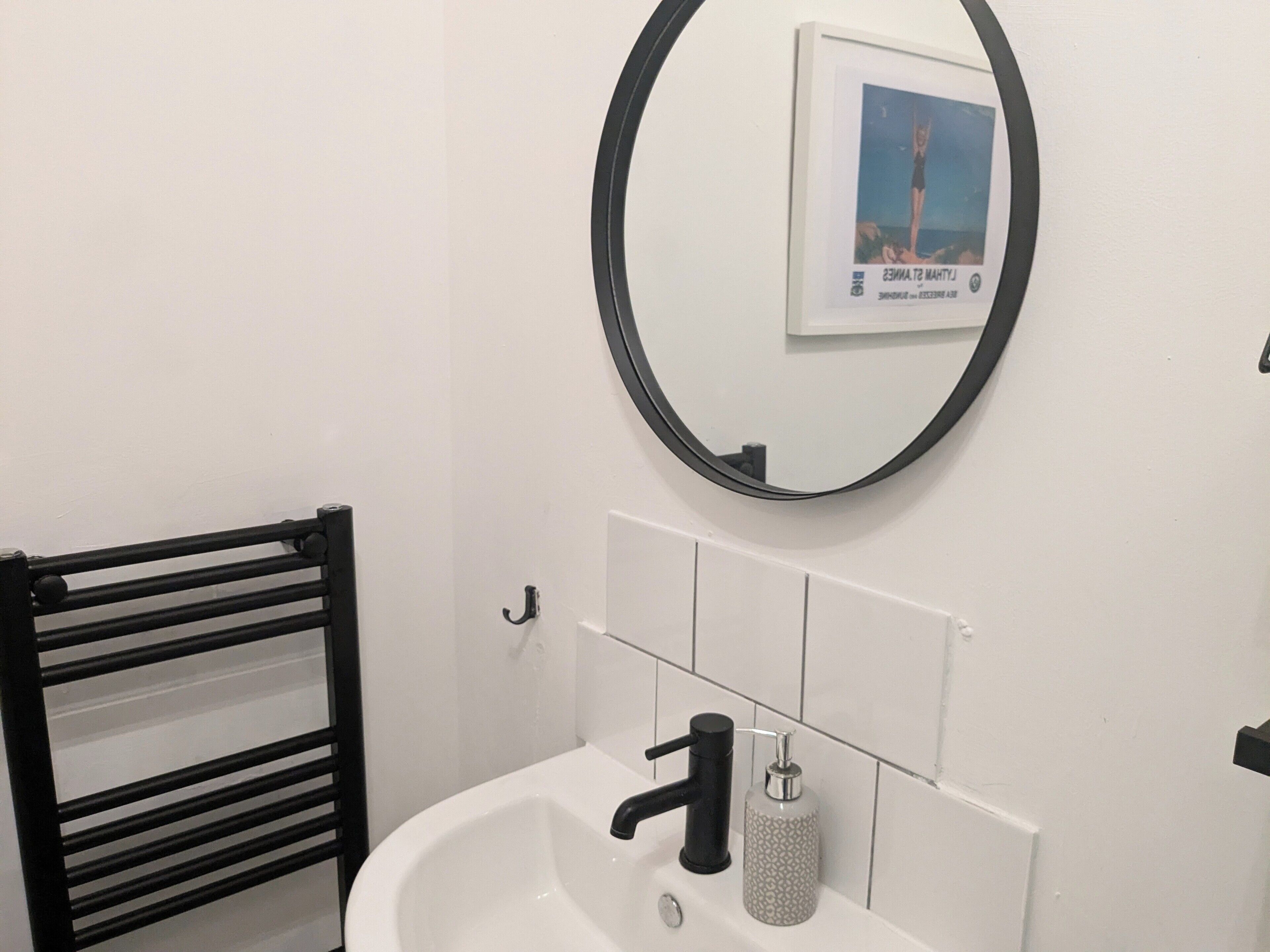 Une salle de bain avec un lavabo blanc, un robinet noir et un miroir rond orné d’un cadre contenant une image.