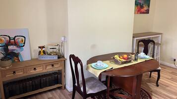 A dining area with a wooden table, chairs, a fruit basket, and a 'WELCOME' sign on a shelf.