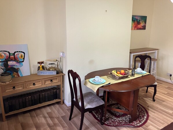Comedor con mesa de madera, sillas, cesta de frutas y un cartel que dice 'WELCOME' en una estantería.