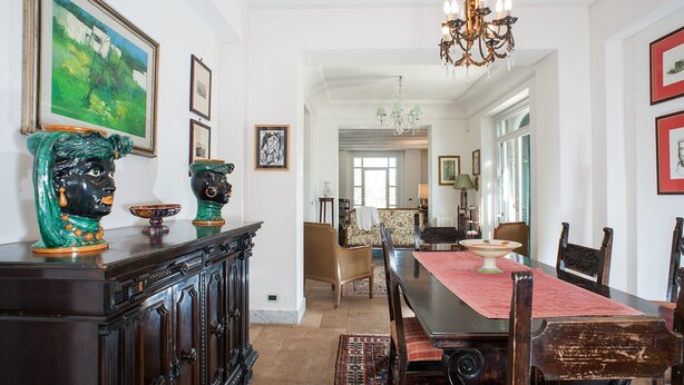 Ein Essbereich mit einem langen Tisch, Stühlen und einer Chandelüstere. An den Wänden hängen gerahmte Bilder und ein Schrank mit dekorativen Gegenständen.