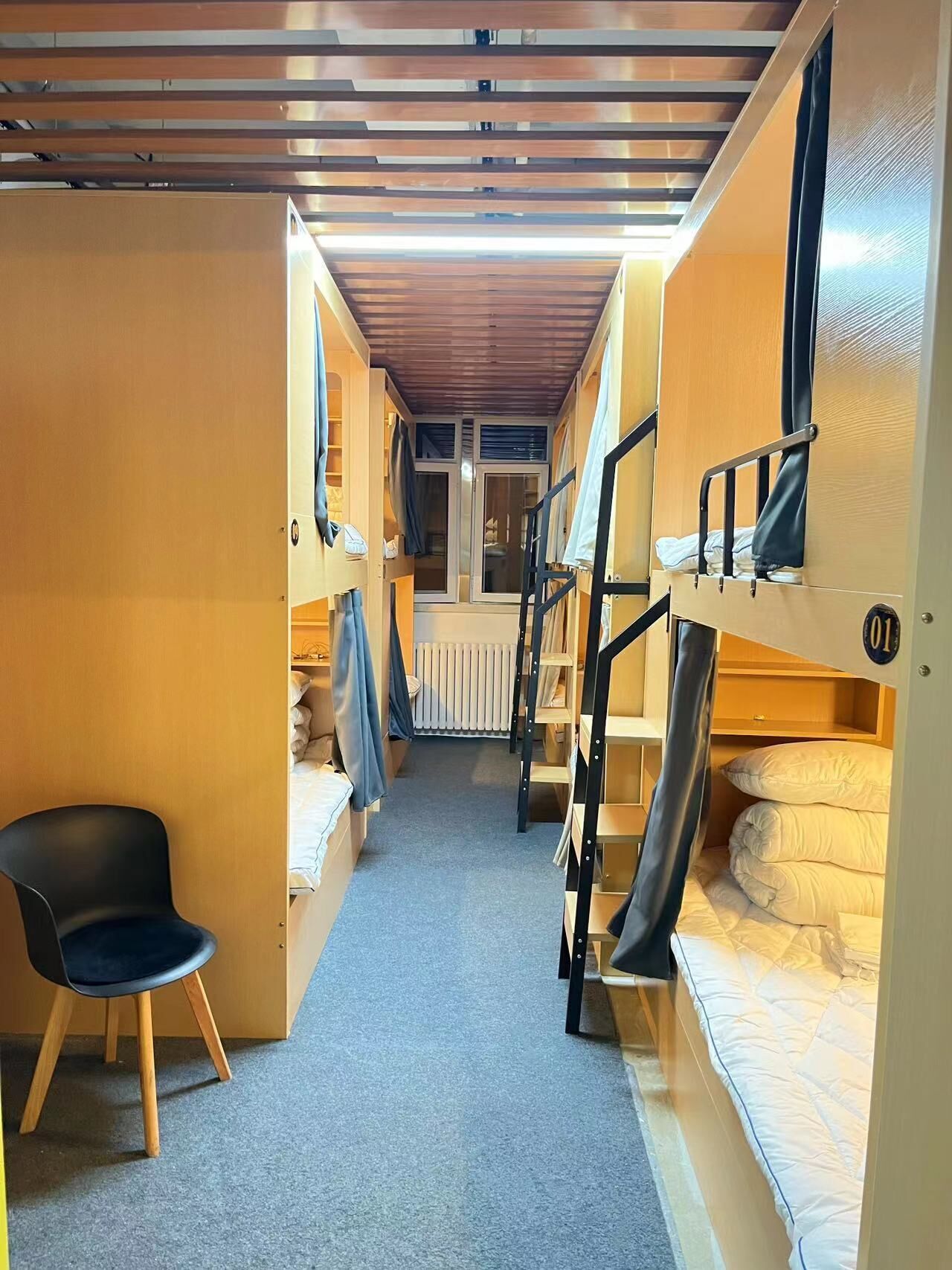 A narrow corridor with bunk beds on both sides, a small seating area with a chair, and a window at the end.