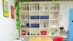 A room with a wooden table and four chairs, a bookshelf filled with books, and a wall with framed pictures.