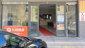 A hotel entrance with a signboard, glass doors, a red carpet, and a digital display inside.