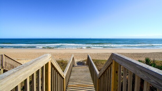 Un paseo marítimo de madera que lleva a una playa bajo un cielo azul despejado.
