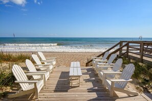 Une terrasse en bois avec des chaises Adirondack blanches et une table, donnant sur une plage où l’on peut admirer les vagues et un ciel dégagé.