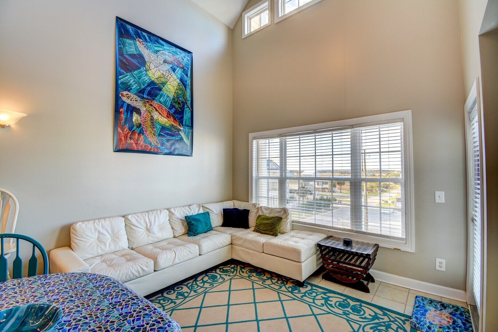 A cozy living room with a white sofa, a colorful abstract painting, and a large window with blinds.