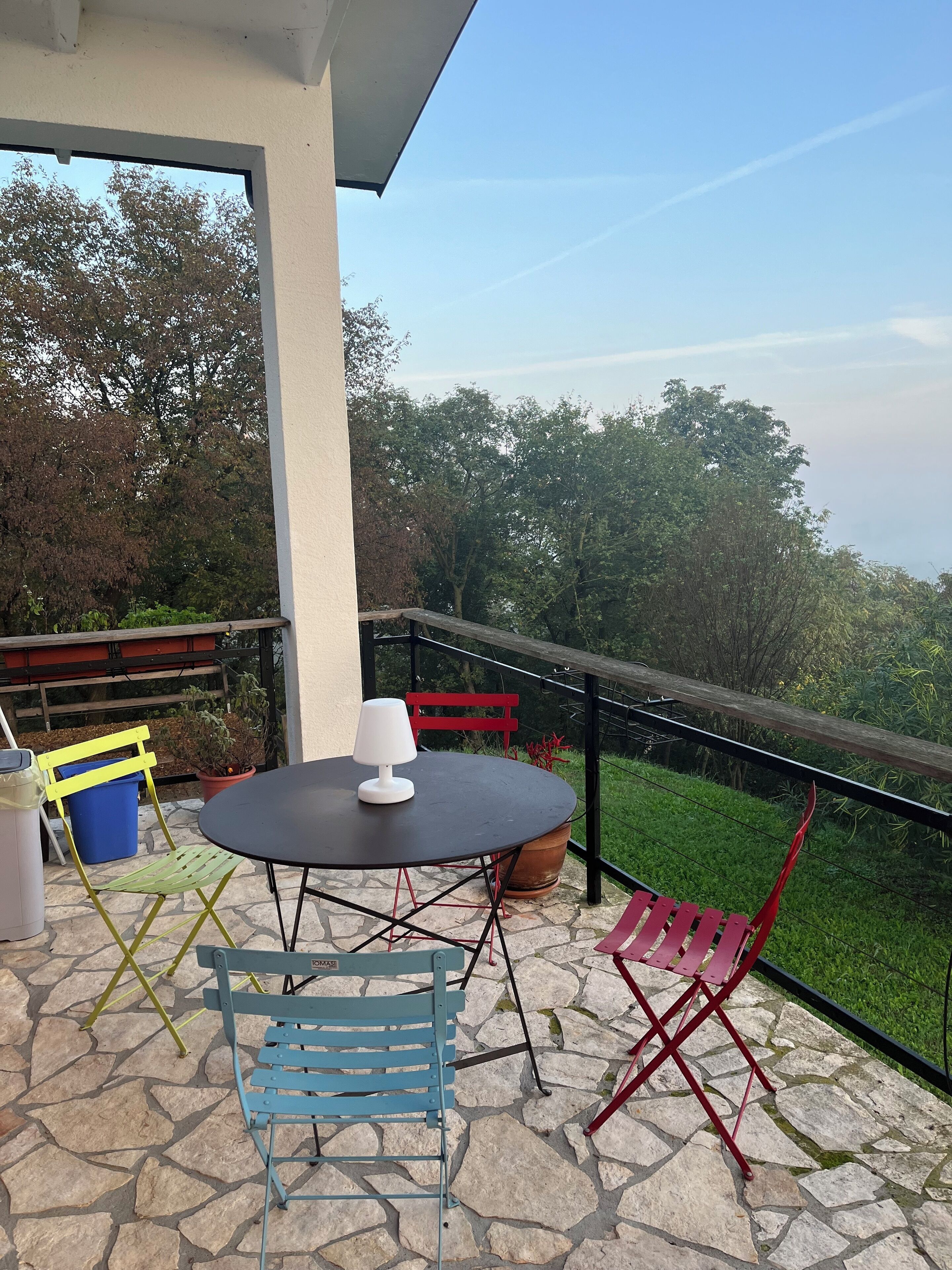 A patio with a round table, two chairs, a potted plant, and a view of trees and grass.