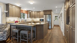 A modern kitchen with wooden cabinets, a stainless steel refrigerator, and a bar area with stools. - Peaceful Riverfront Hideaway | 4BR/3BA | Wildlife, Views & Deep Water Dock (Edgecomb)