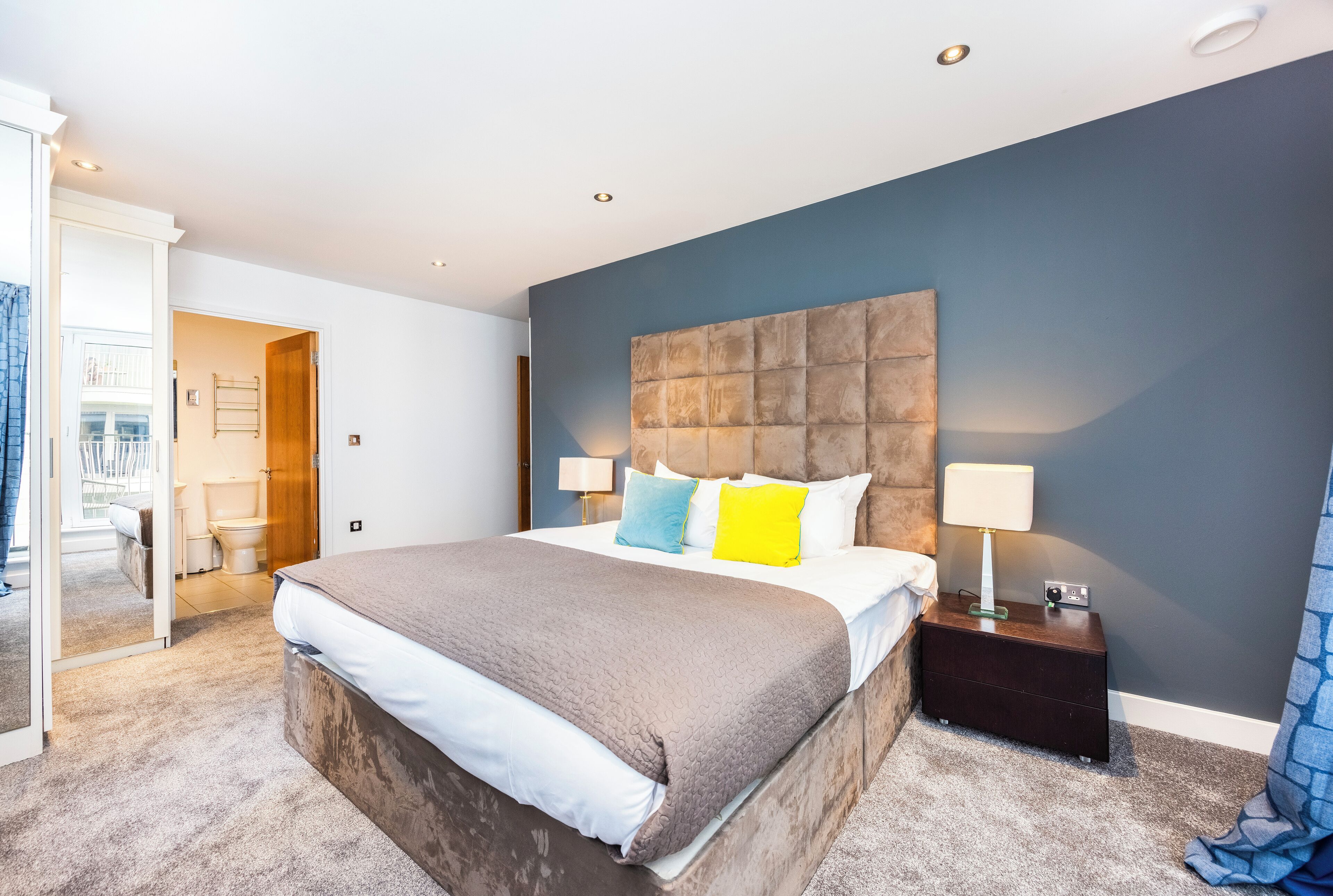 A modern bedroom with a large bed, a nightstand with a lamp, and a view of the outside balcony.