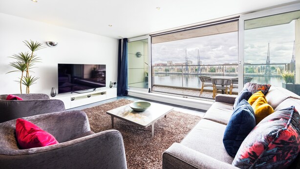 A modern living room with a large TV, comfortable seating, a coffee table, and a view of a cityscape through large windows. - Koks Mews 2 Bed 2 Bath Apartment - ExCeL (London)