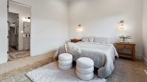 Un dormitorio con una cama, dos mesitas de noche, una mesita auxiliar, una planta en maceta y un vano que da a un baño.