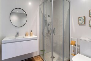 Un baño moderno con ducha envuelta en vidrio, un espejo redondo, un lavabo blanco y un inodoro con dispensador de papel higiénico de madera.