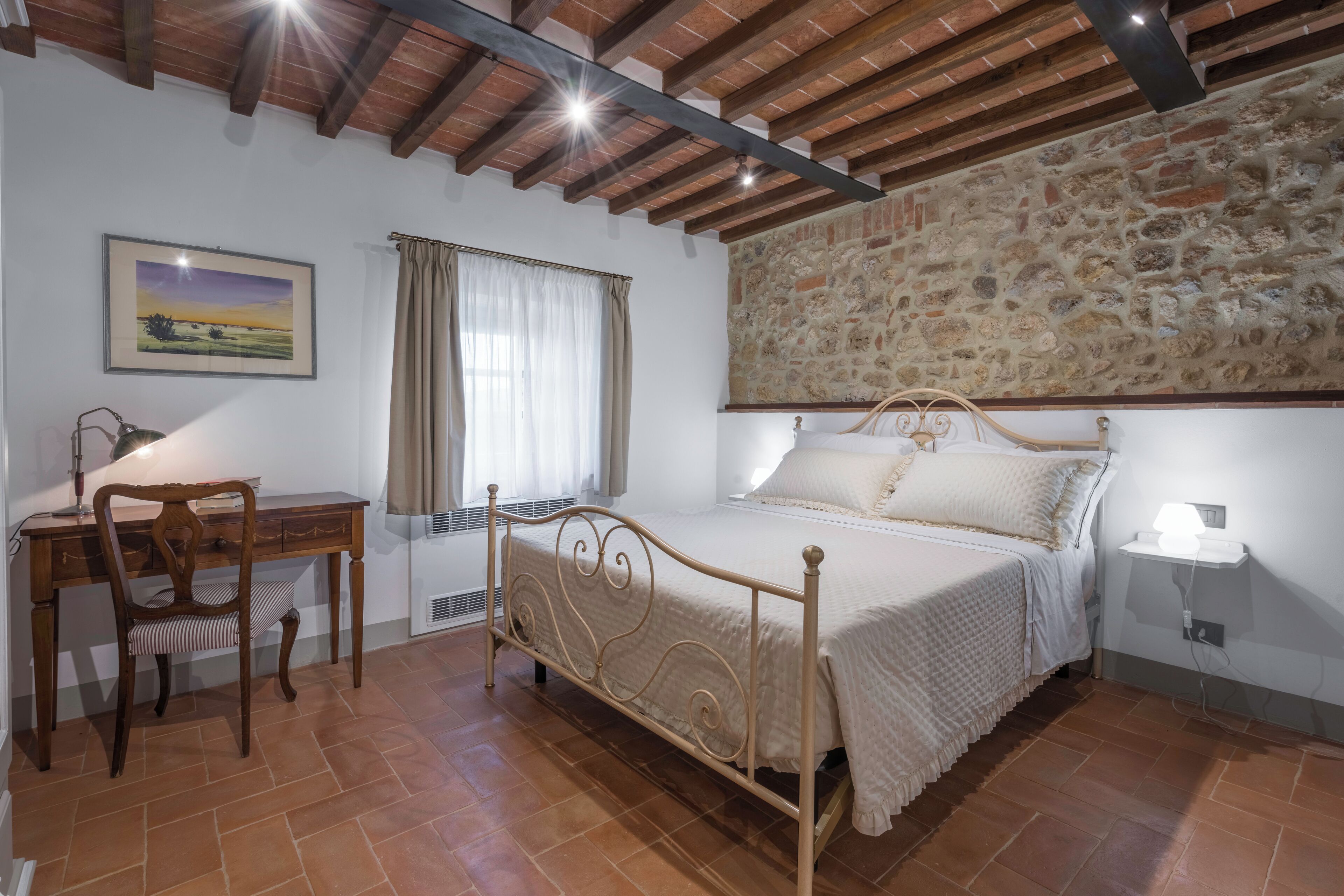 Una camera da letto con un letto, una scrivania, una sedia, una finestra con tende e un muro in pietra.