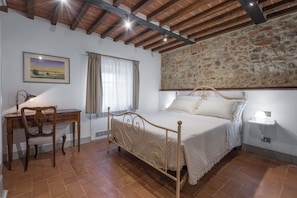 A bedroom with a bed, a desk, a chair, a window with curtains, and a stone wall.