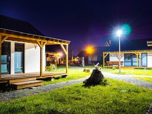 A well-lit outdoor area with a wooden deck, a covered seating area, and a building with a number sign '6'. - 2-bed Holiday Homes With Pool (Postomino)