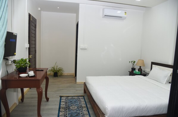 A hotel room with a bed, a desk with a plant, a small table with a plant, an air conditioning unit, and a nightstand with a lamp. - Arambol Sweet Lake Resort (Arambol)