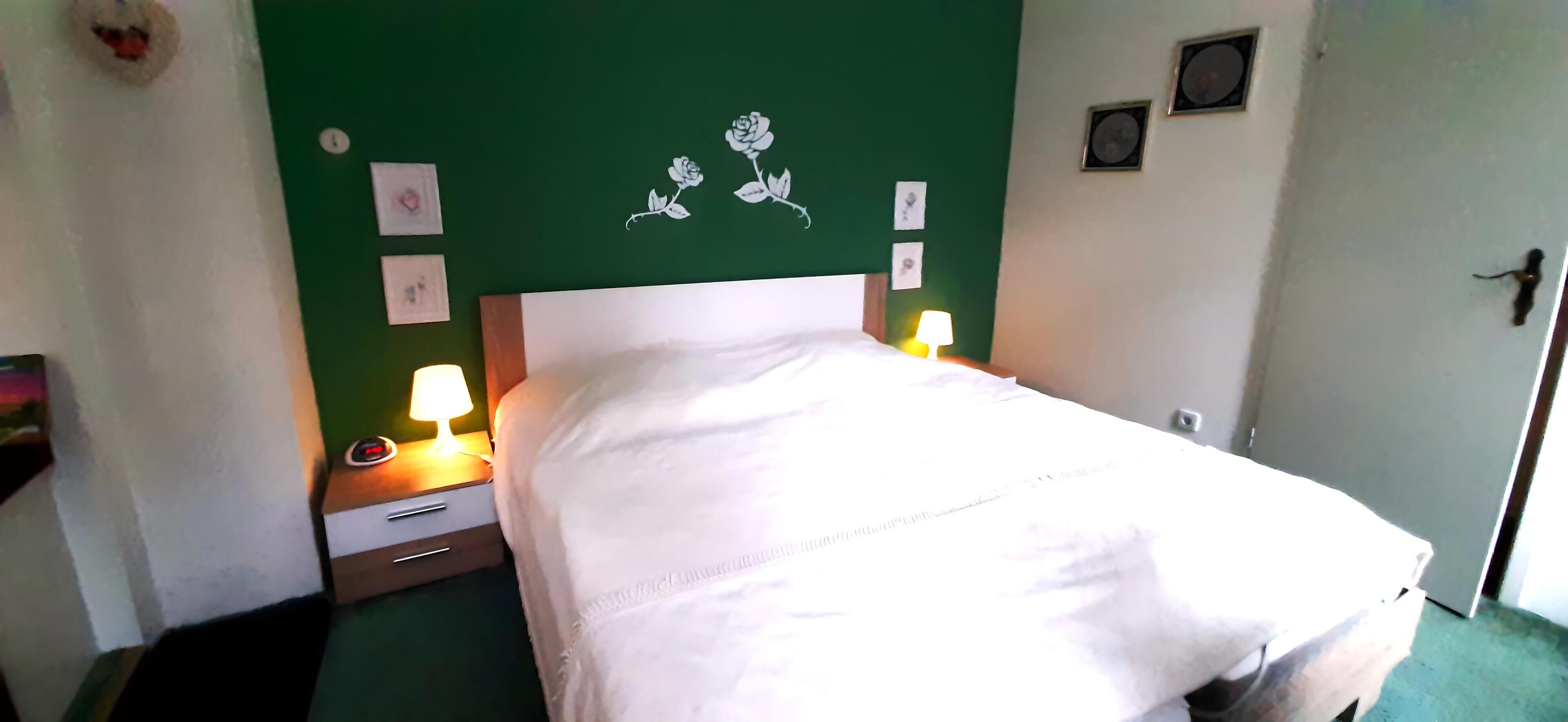 Una cama bien hecha en una habitación con paredes verdes, un cabecero blanco y una mesita de noche con lámpara.