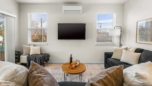 Una sala de estar con un televisor de pantalla plana, una mesa de centro redonda de madera con aperitivos, un sofá gris y una ventana con persianas.