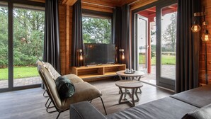 A modern living room with a large TV, wooden furniture, and a glass door leading outside. - Lodge by the Lake in Arcen's Tranquil Nature (Arcen)