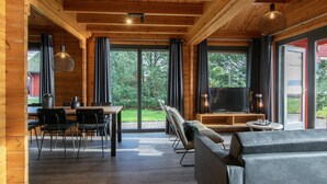 A spacious living area with a wooden ceiling, large windows, a sofa, a TV, and a dining table with chairs. - Lodge by the Lake in Arcen's Tranquil Nature (Arcen)