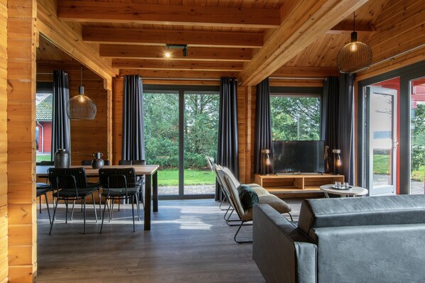 A spacious living area with a wooden ceiling, large windows, a sofa, a TV, and a dining table with chairs. - Lodge by the Lake in Arcen's Tranquil Nature (Arcen)