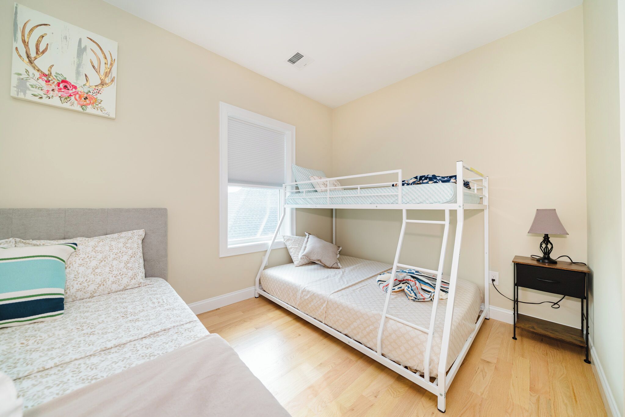A bedroom with a bunk bed, a single bed, a nightstand, a lamp, and a window with blinds.
