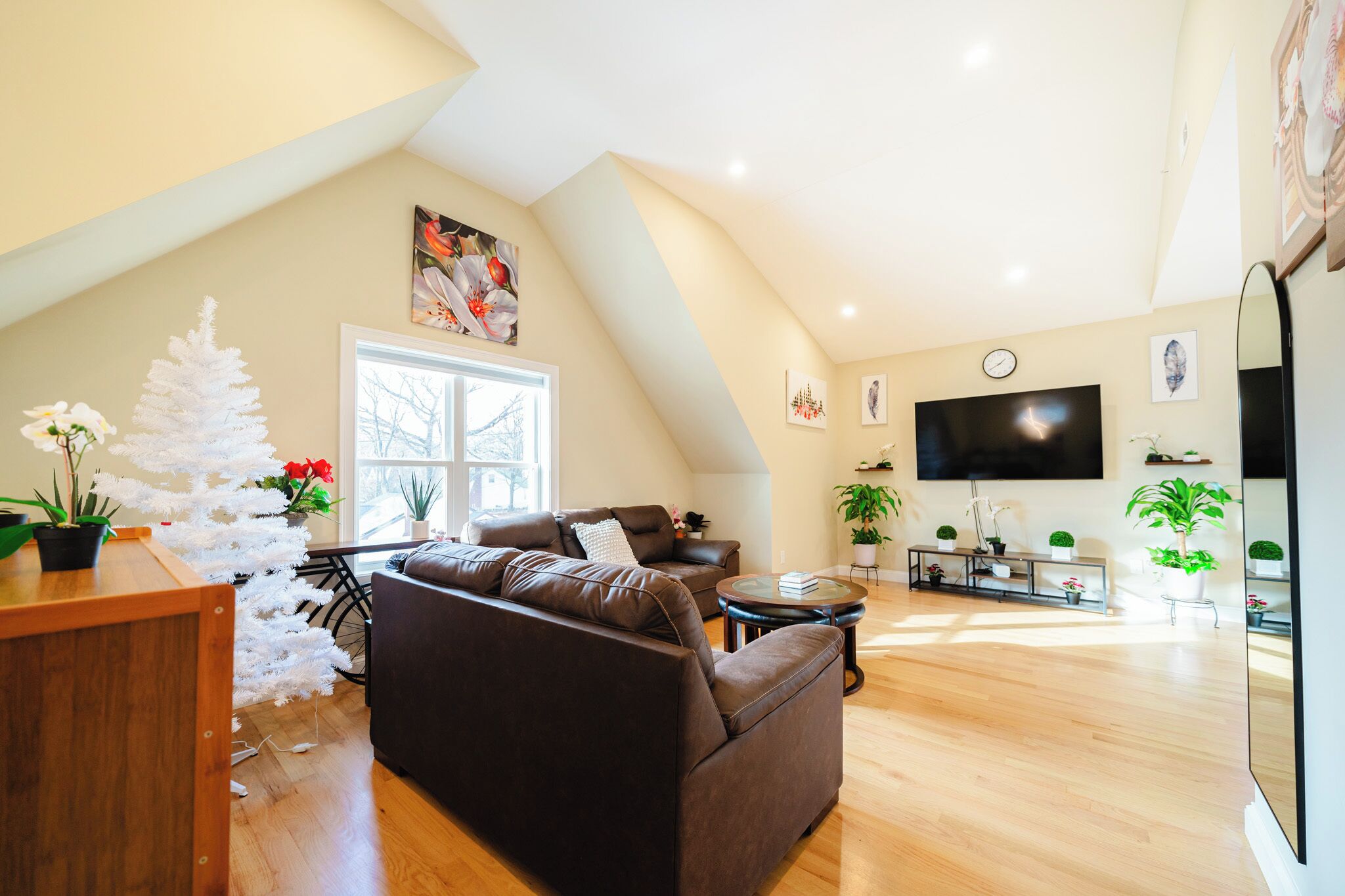 A modern living room with a brown leather sofa, a white Christmas tree, and a large flat-screen TV mounted on the wall.