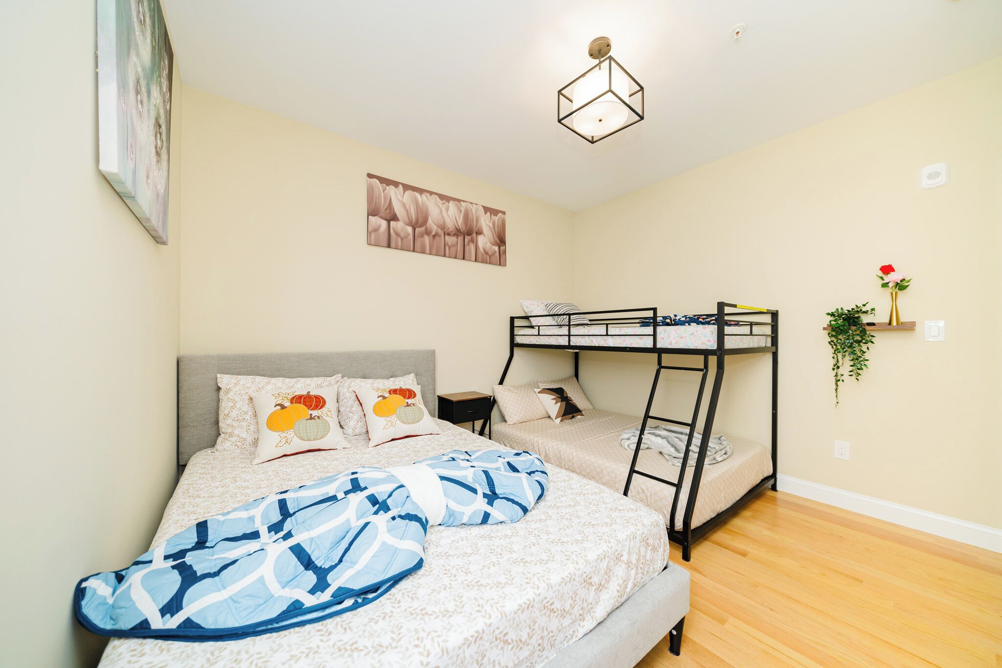 A bedroom with a bunk bed, a single bed with patterned bedding, a ceiling light fixture, and a wall-mounted plant.