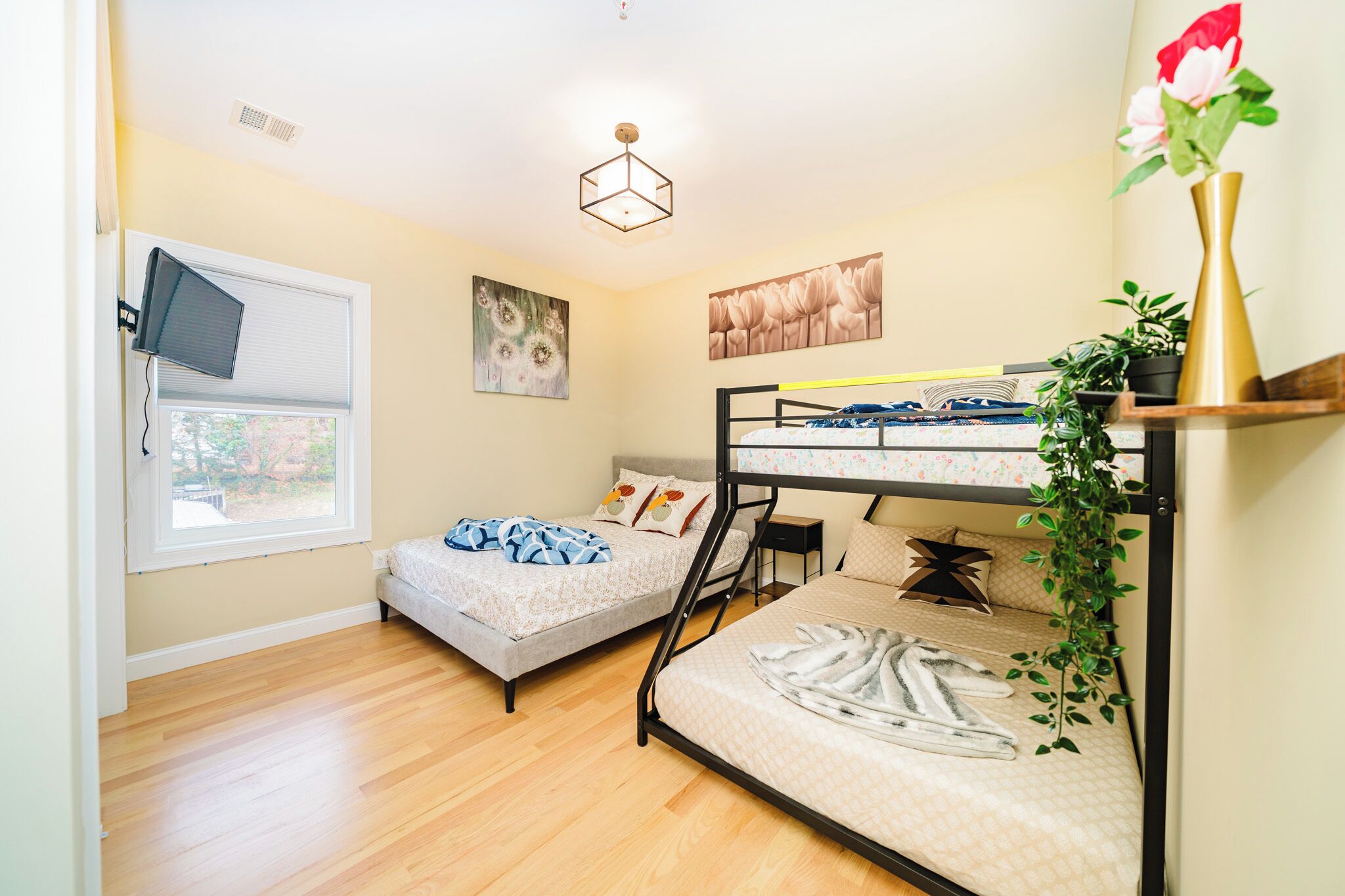 A room with two bunk beds, a window with blinds, a desk, a chair, a plant, and artwork on the walls.