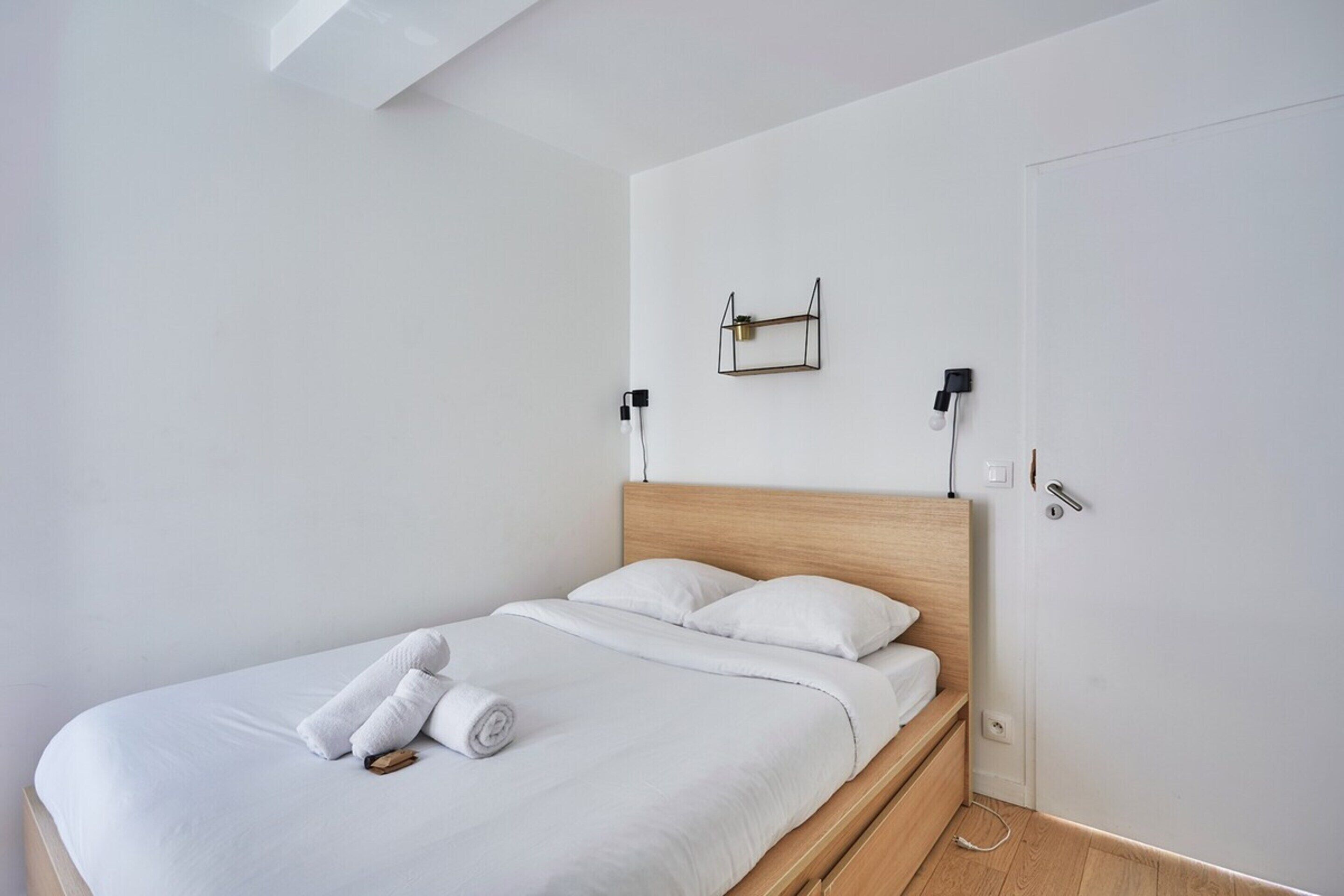 Un lit bien fait, avec du linge de lit blanc et une serviette pliée, une tête de lit en bois et une petite étagère sur laquelle est posée une lampe.
