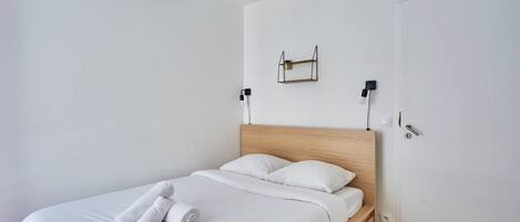 Un lit bien fait, avec du linge de lit blanc et une serviette pliée, une tête de lit en bois et une petite étagère sur laquelle est posée une lampe.