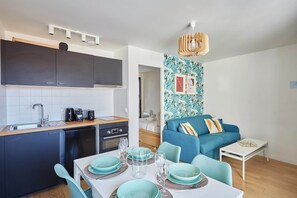 A compact living space with a kitchenette, dining area, and a blue sofa with cushions. - Modern Apartment Boulogne Facing "les Passages" (Boulogne-Billancourt)