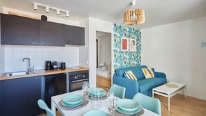 A compact living space with a kitchenette, dining area, and a living room with a blue sofa and patterned wallpaper. - Modern Apartment Boulogne Facing "les Passages" (Boulogne-Billancourt)