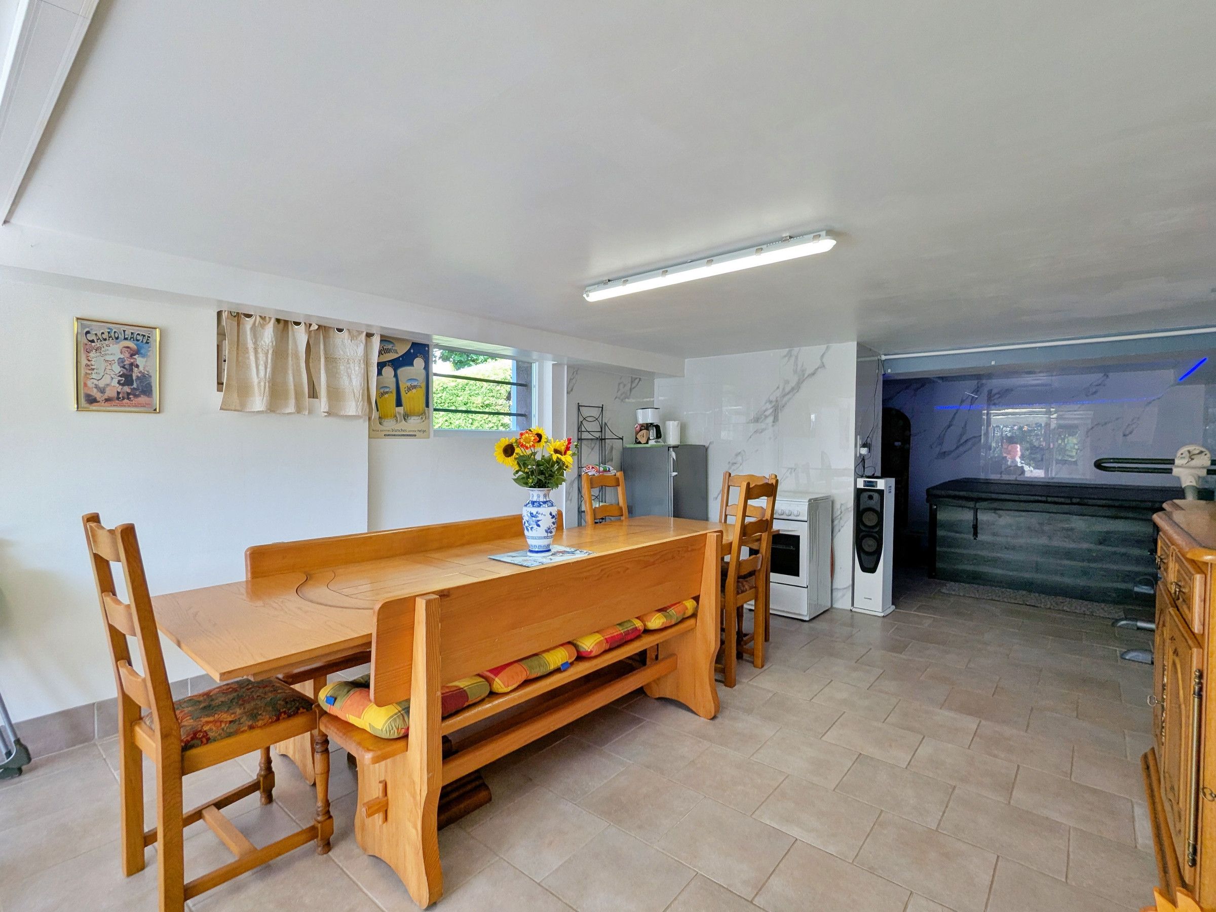 Een eetgedeelte met een houten tafel en stoelen, een vaas met bloemen, een keuken met apparatuur, en uitzicht op de omgeving.