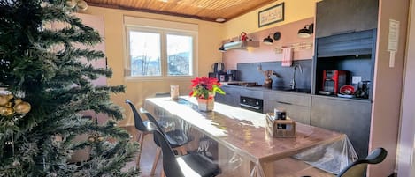 Un espace repas avec un plafond en bois, un sapin de Noël, une table avec des chaises et un coin cuisine équipé d’appareils électroménagers.