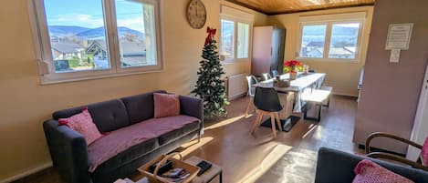 A cozy living room with a sofa, coffee table, and a dining area with a table and chairs. There is a Christmas tree and a clock on the wall.