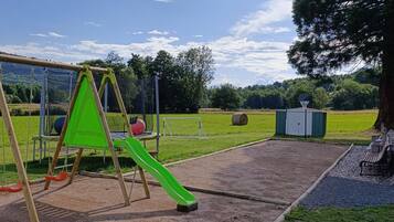 Une aire de jeux avec des balançoires et un toboggan, entourée d’un champ d’herbe et d’arbres.