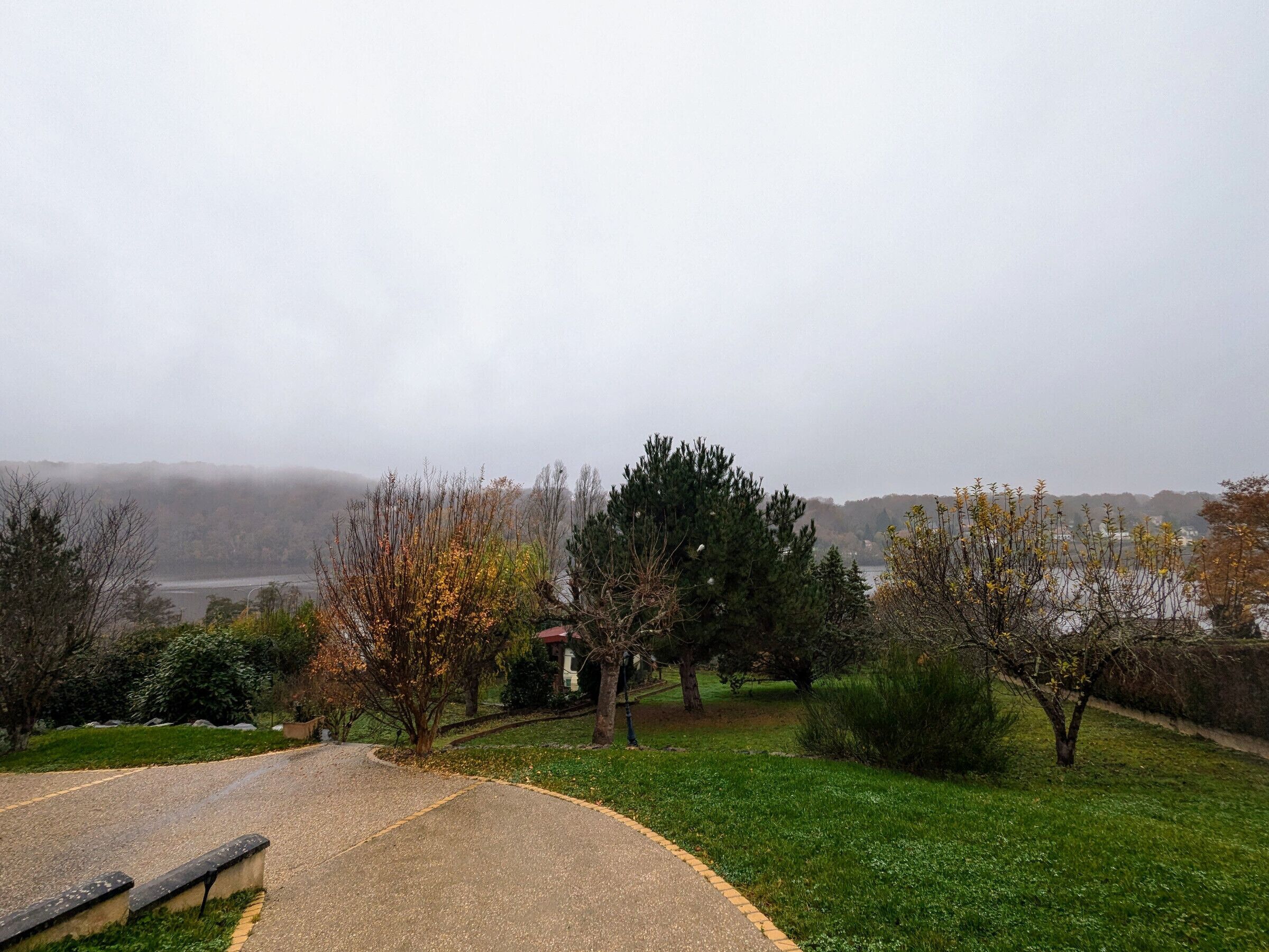 Un parc avec un sentier dallé, des arbres et une vue sur un lac et des collines lointaines.