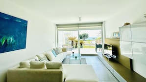 Una sala de estar moderna con un sofá, una mesa de centro de vidrio y un televisor. Hay un ventanal que ofrece vistas al exterior.