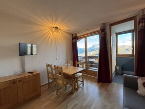 Ein Essbereich mit Holztisch und Stühlen, einem Fernseher auf einem Schrank und Blick auf eine Berglandschaft durch große Fenster.
