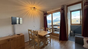 Un coin repas avec une table et des chaises en bois, un téléviseur posé sur un meuble, et une vue sur un paysage montagneux grâce à de grandes fenêtres.