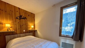 Une chambre à coucher avec une tête de lit en bois, deux lampes de chevet, un lit recouvert d’une couette blanche et une fenêtre donnant sur un paysage enneigé.