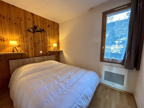 Een slaapkamer met een houten hoofdbord, twee bedlampen, een bed met een witte dekbedovertrek en een raam met uitzicht op sneeuw.