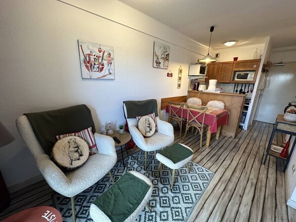 Una sala de estar acogedora con dos sillones, una mesa de comedor con sillas, un tapete estampado y una zona de cocina con armarios de madera y un microondas.