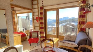 A cozy room with a wooden table set for dining, a sofa, and a chair. A ladder is visible against the wall, and a window offers a view of mountains. - Studio Les Saisies 4 people with balcony (Hauteluce)