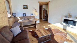 En hyggelig stue med en brun lædersofa, et træsofabord og en kohudes måtte. Der er et spisebord med stole, en træbænk og et bogreol i baggrunden.