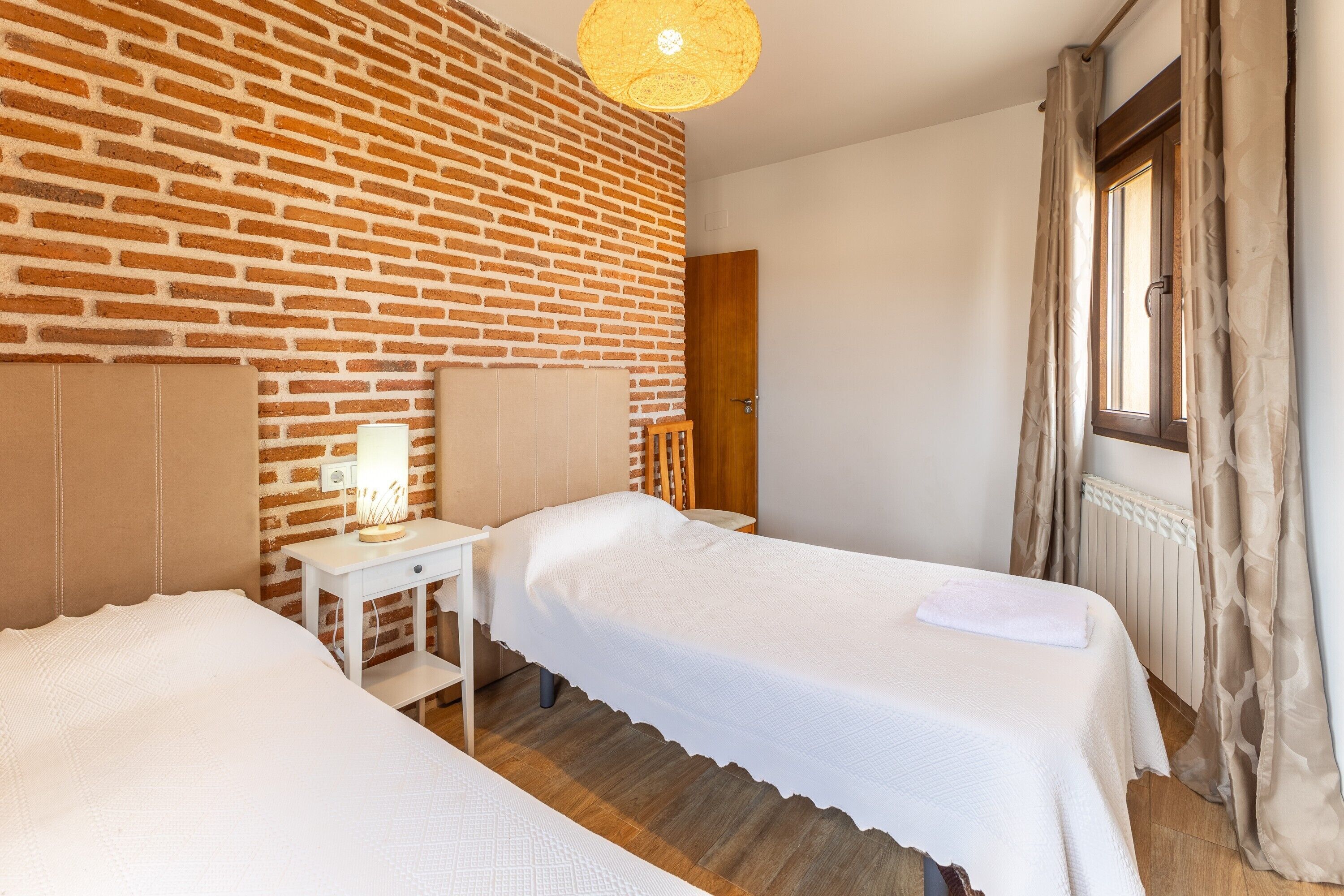 A room with two beds, a brick wall, a wooden floor, a window with curtains, and a hanging light fixture.