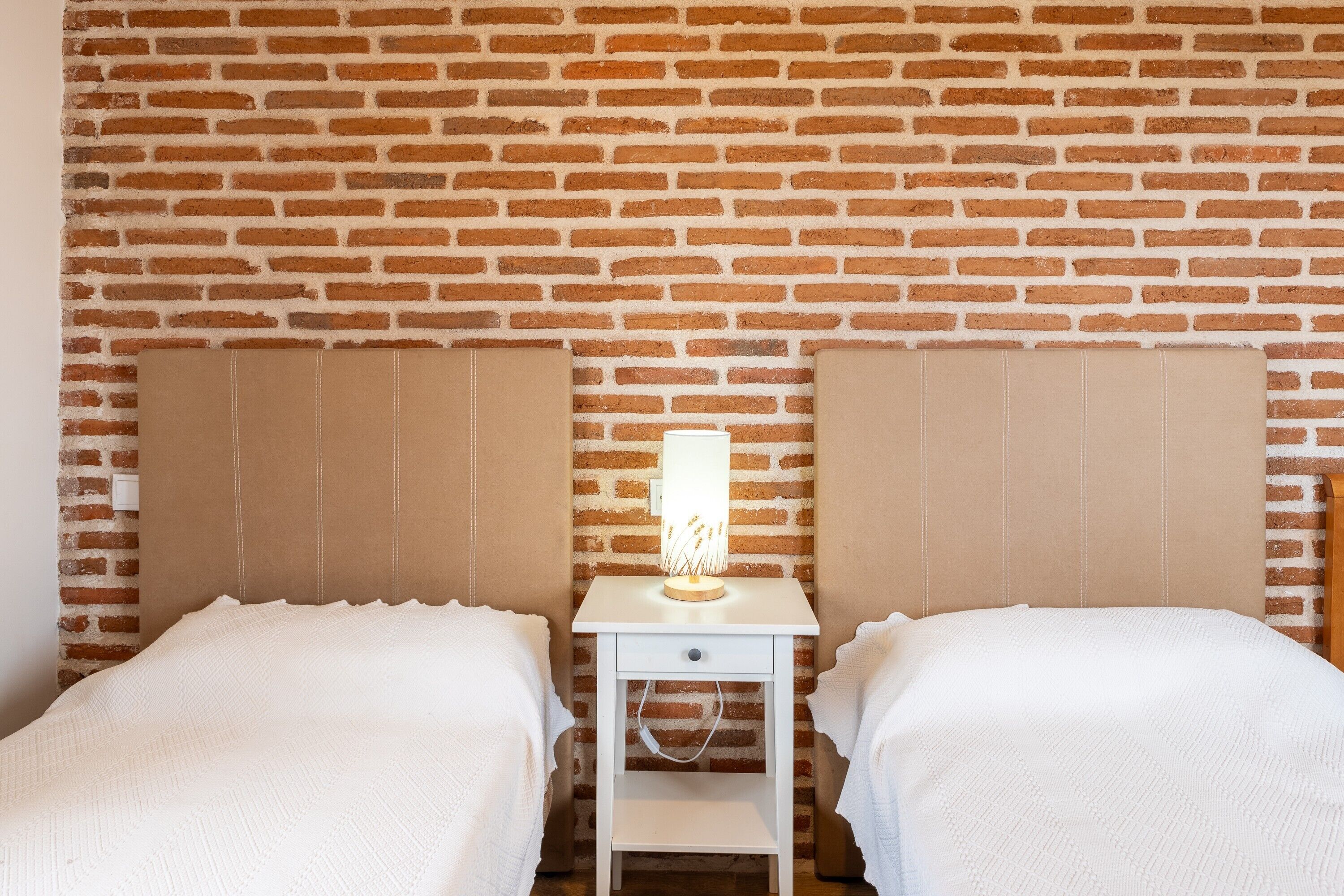 A room with a brick wall, two beds with white bedding, a nightstand with a lamp, and a drawer.
