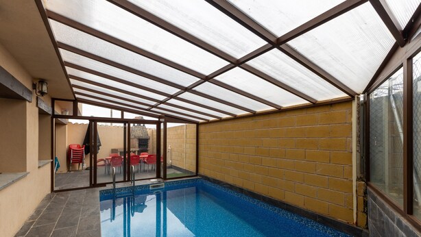 A covered indoor swimming pool with a glass wall, a brick wall, and a glass door leading to an outdoor area with red chairs. - Country House 'Camino De Avila' with Mountain View, Private Pool and Wi-Fi (Mediana De Voltoya)