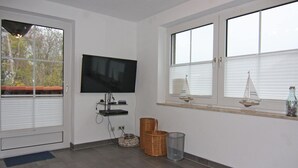 Una habitación con una televisión de pantalla plana en la pared, una ventana con persianas, un estante pequeño con adornos y una cesta de mimbre en el suelo.